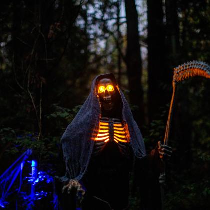 Halloween skeleton with glowing eyes holds a scythe in a dark forest.