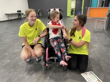 Staff members kneel beside a child seated in a wheelchair inside a community centre room