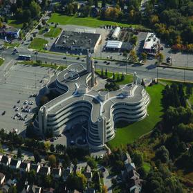Aerial view of a large, curvy building with a clock tower surrounded by greenery and roads.